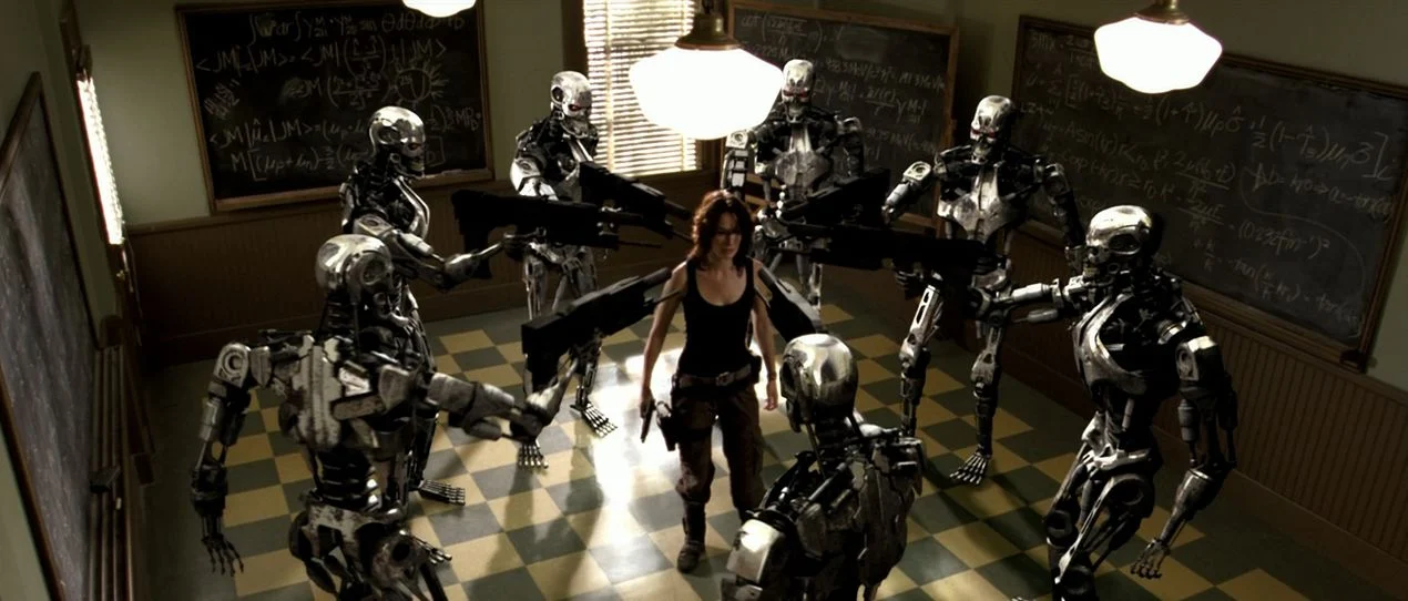 Terminator-Kroniki-Sary-Connor-2 | film.org.pl Woman in a black tank top stands in the center, surrounded by chrome robots with weapons in a classroom with chalkboards and a checkered floor.