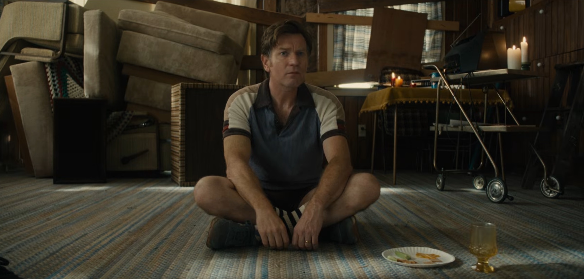 Man sits cross-legged on a striped rug in a cluttered basement, surrounded by furniture and a candle-lit setup near a table with a walker nearby.