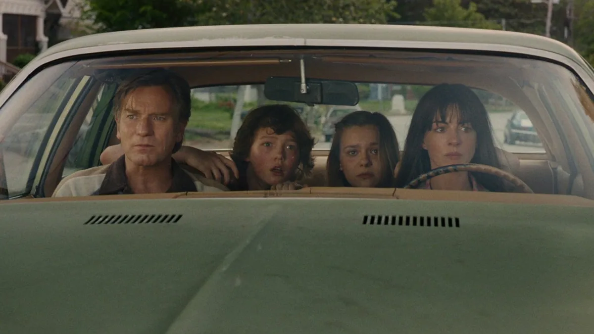 Man and woman ride in a green car with two children in the back, all looking ahead with concerned expressions as they drive.