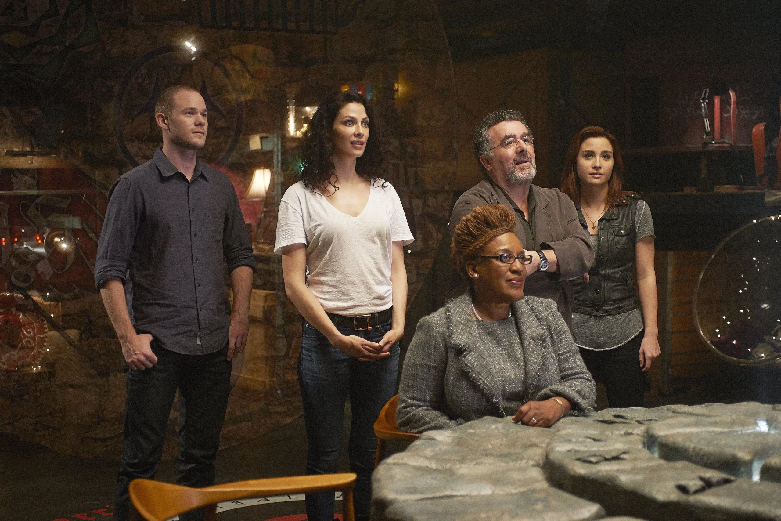 Five adults stand and sit around a stone table in a dim gallery with rocky walls and glass partitions, looking toward something off-camera.