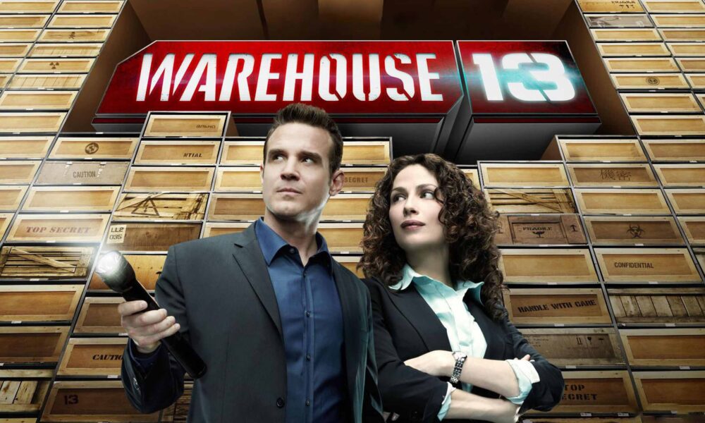 Two businesspeople in suits stand in front of a large red 'WAREHOUSE 13' sign amid stacked wooden crates in a warehouse setting, looking off to the side.