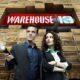 Two businesspeople in suits stand in front of a large red 'WAREHOUSE 13' sign amid stacked wooden crates in a warehouse setting, looking off to the side.