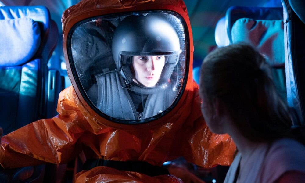 Person in an orange hazmat suit and helmet sits in a bus, speaking with a passenger seated nearby.