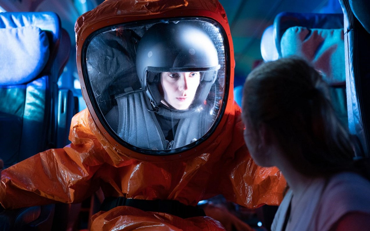 Person in an orange hazmat suit and helmet sits in a bus, speaking with a passenger seated nearby.