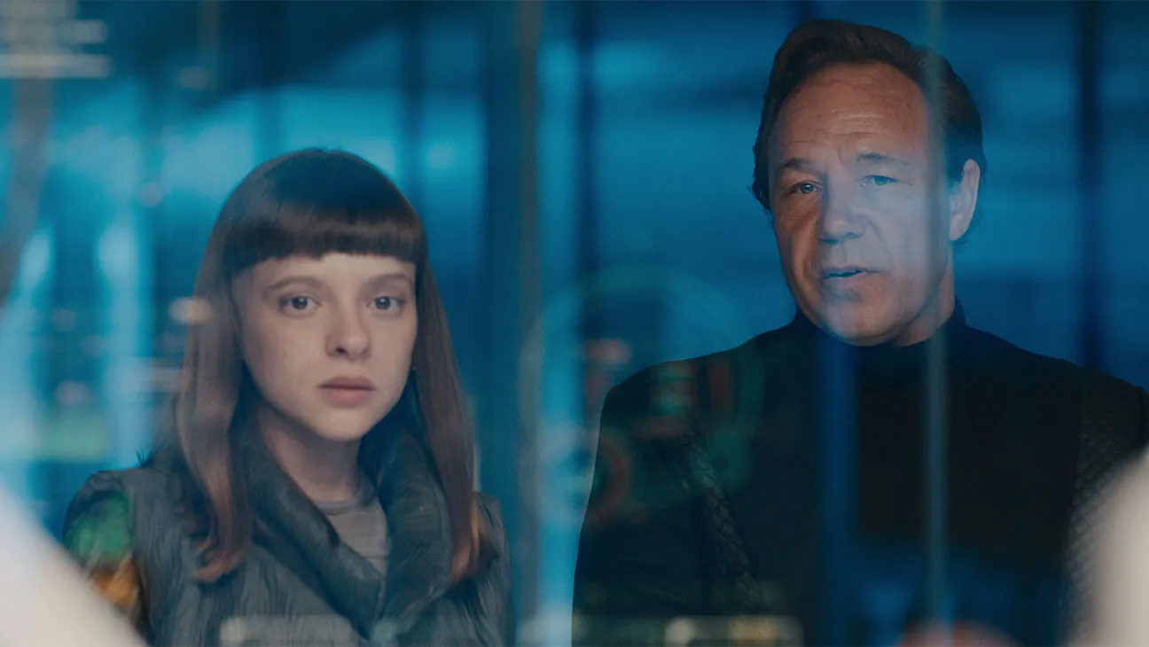 Two people stand behind a glass barrier in a blue-lit, high-tech setting, looking intently at something off-camera.