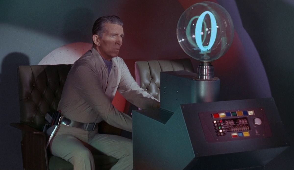 Man in a beige sci-fi uniform seated at a retro spaceship console with a glowing glass orb on top, working the controls.