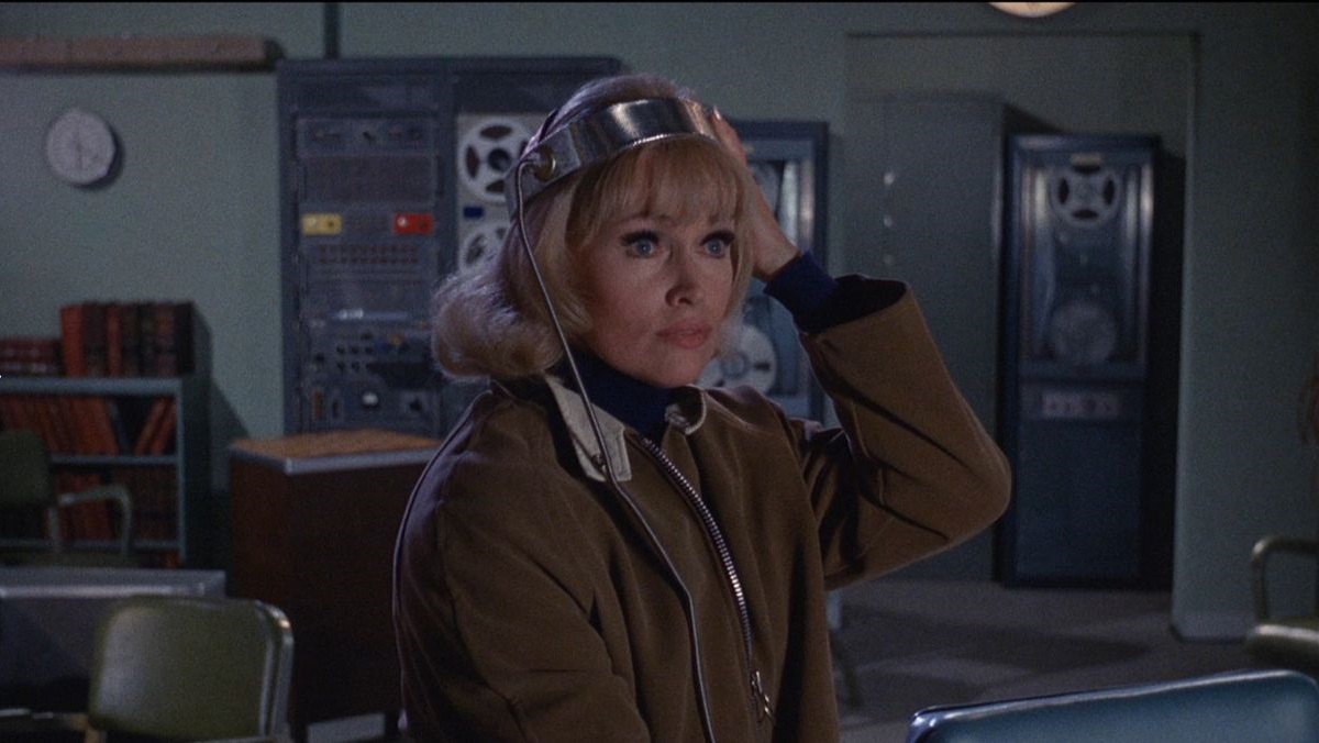 Woman in a brown jacket wearing a headset, in a retro control room with tape machines behind her.