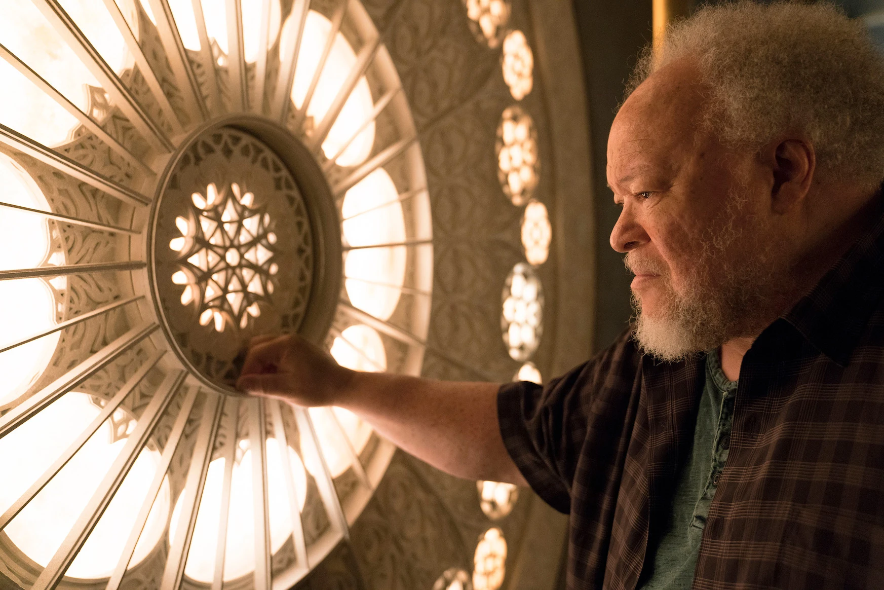 Older man with a gray beard touches a large ornate circular light fixture, glowing warmly in a dim setting, profile view.