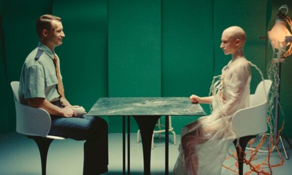 Two people sit at a metal table in a green room: a man in a uniform on the left and a pale, bald figure in a translucent gown connected to cables on the right.