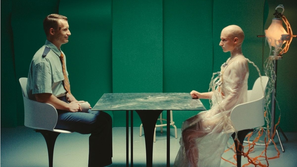 Two people sit at a metal table in a green room: a man in a uniform on the left and a pale, bald figure in a translucent gown connected to cables on the right.