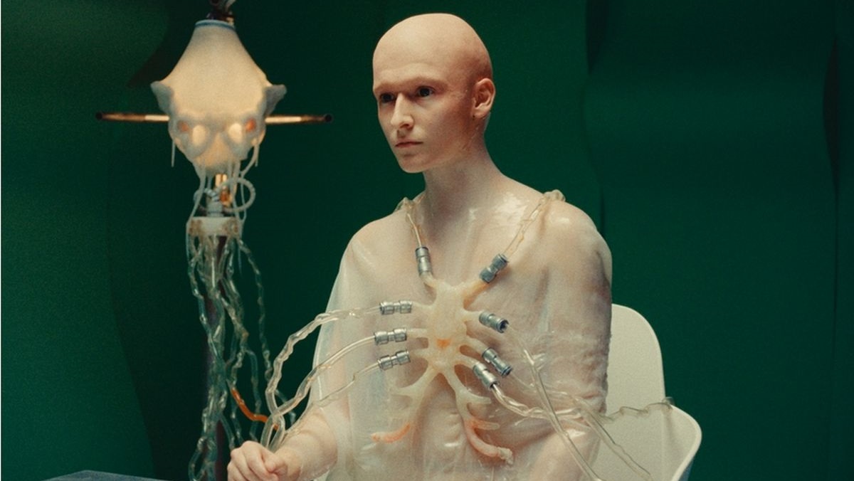 A pale, bald humanoid mannequin connected to clear tubes and metal connectors across its chest in a laboratory-like green room.