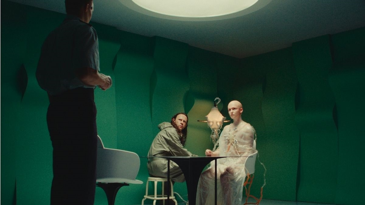Sci‑fi lab scene: a bald, translucent humanoid with visible skeleton sits at a table with two real men in a green-walled room; a standing man faces them from the left.