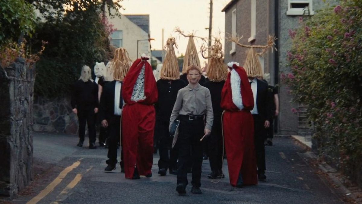 A street parade with performers wearing tall straw hats and red cloaks, led by a person in a beige shirt holding a folder.