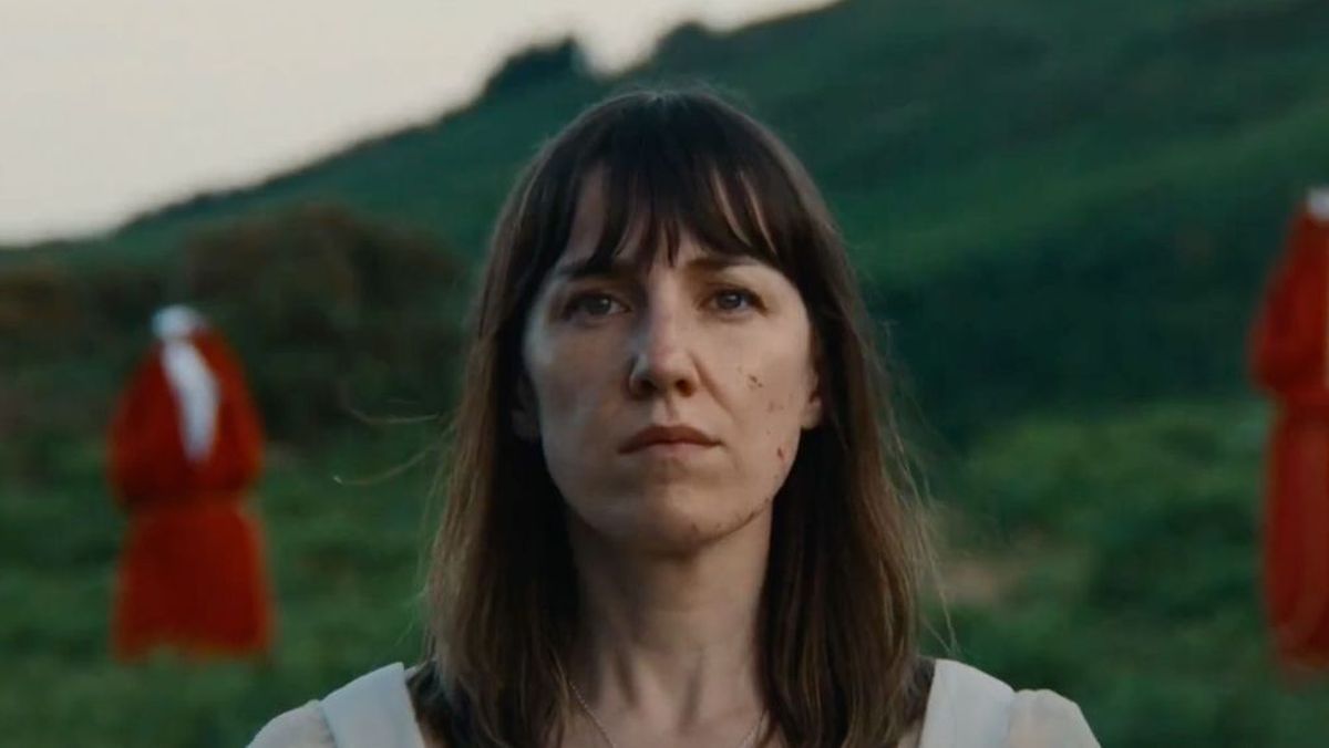 Close-up of a woman with shoulder-length brown hair and bangs, standing outdoors in a grassy landscape with two red shapes in the background.