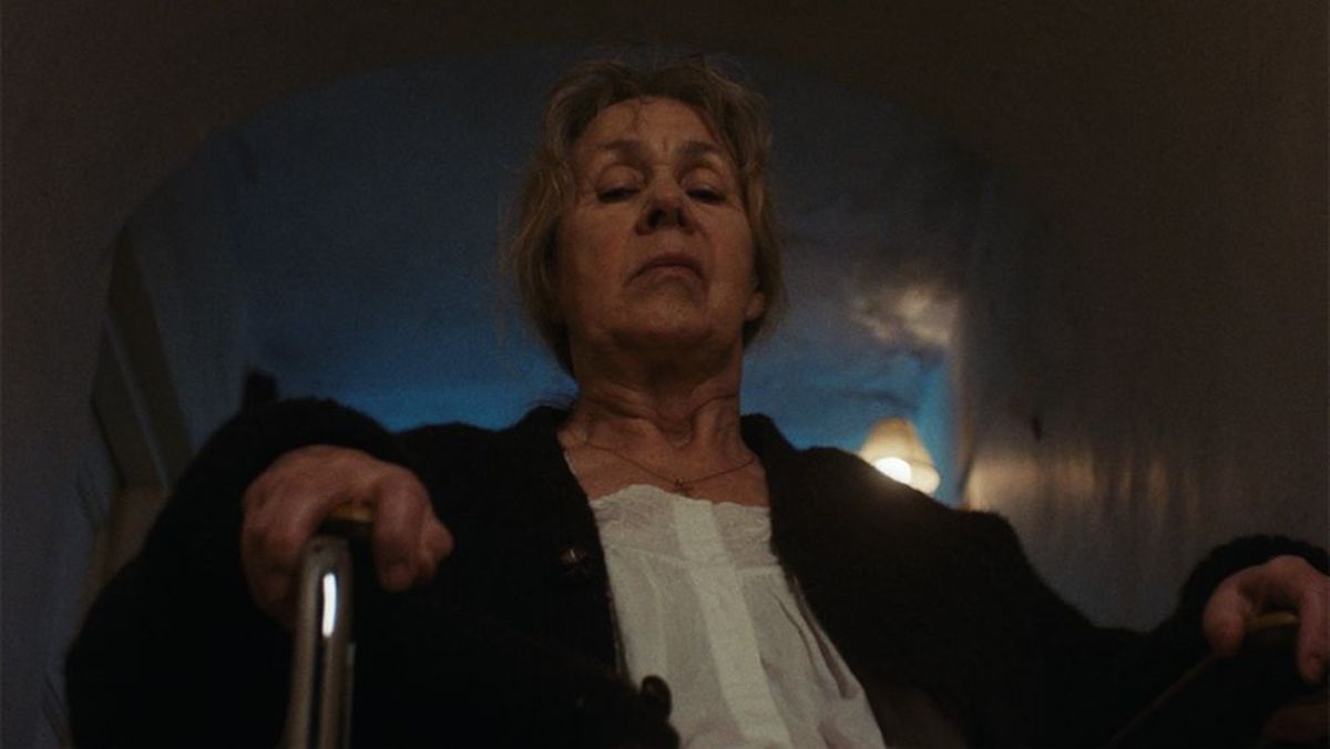 Older woman wearing a white blouse, holding a walker in a dim hallway.”] ,