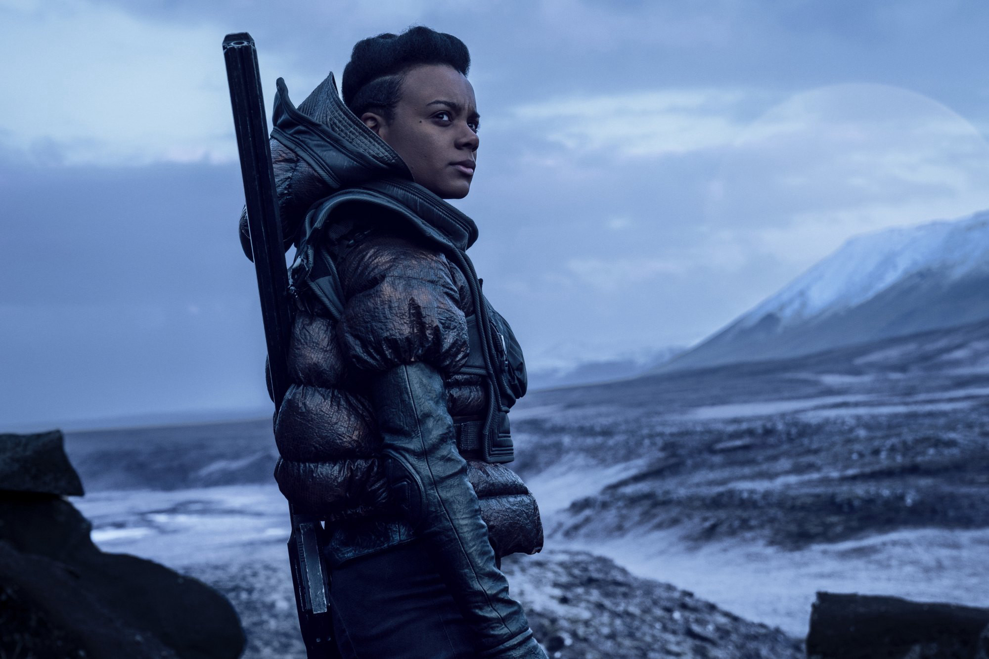Person in a heavy winter jacket with a rifle slung over the shoulder stands on a rocky, snowy shoreline, looking into the distance.