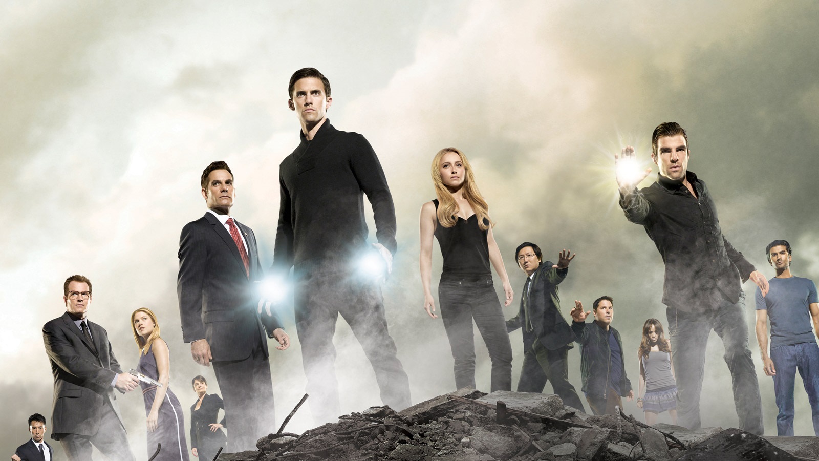 Group of diverse adults standing on rubble in a smoky, cloudy landscape; foreground figure glows with energy in his hand, suggesting powers.