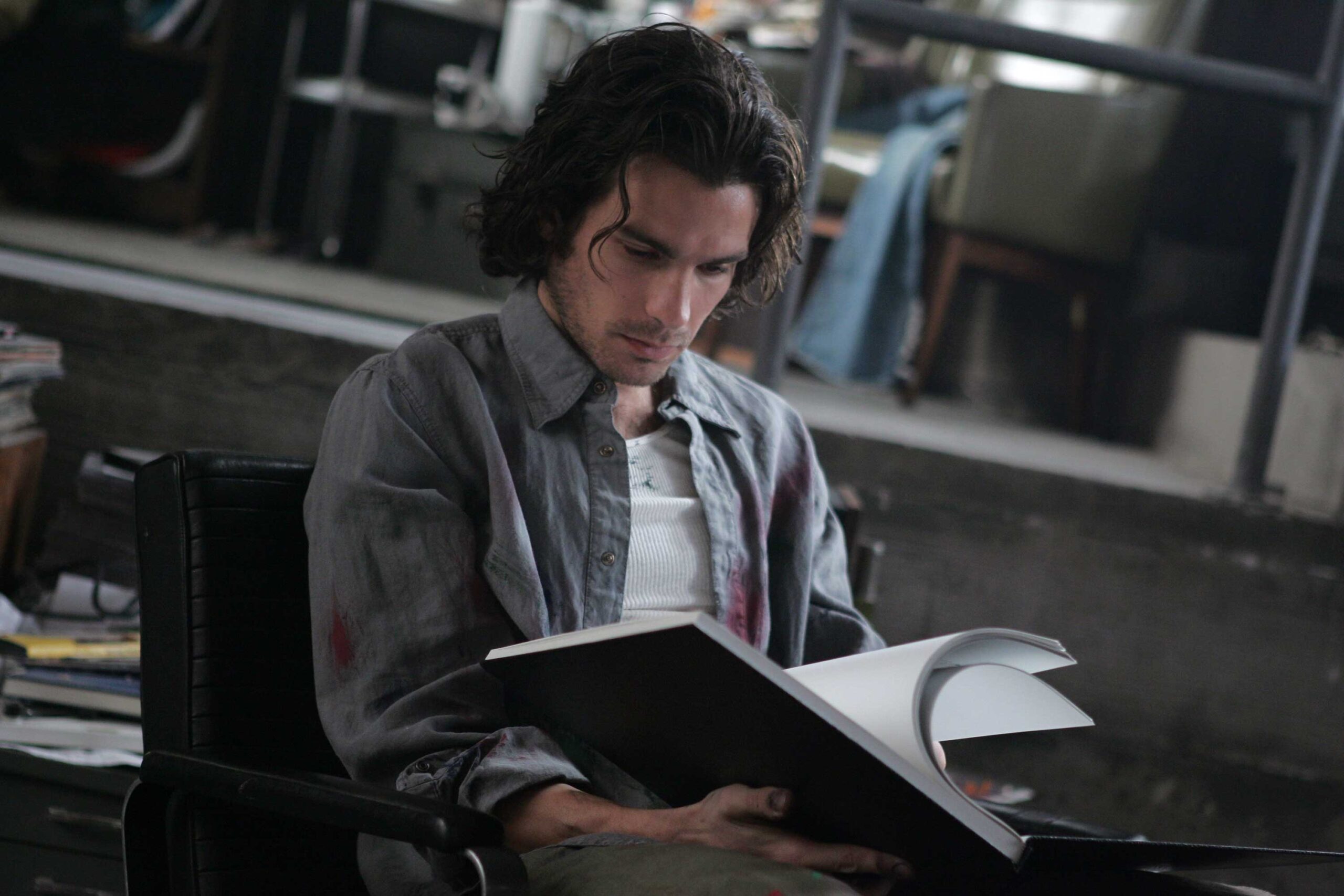 Man in a wheelchair reading an open binder or portfolio in a dim, cluttered workspace.