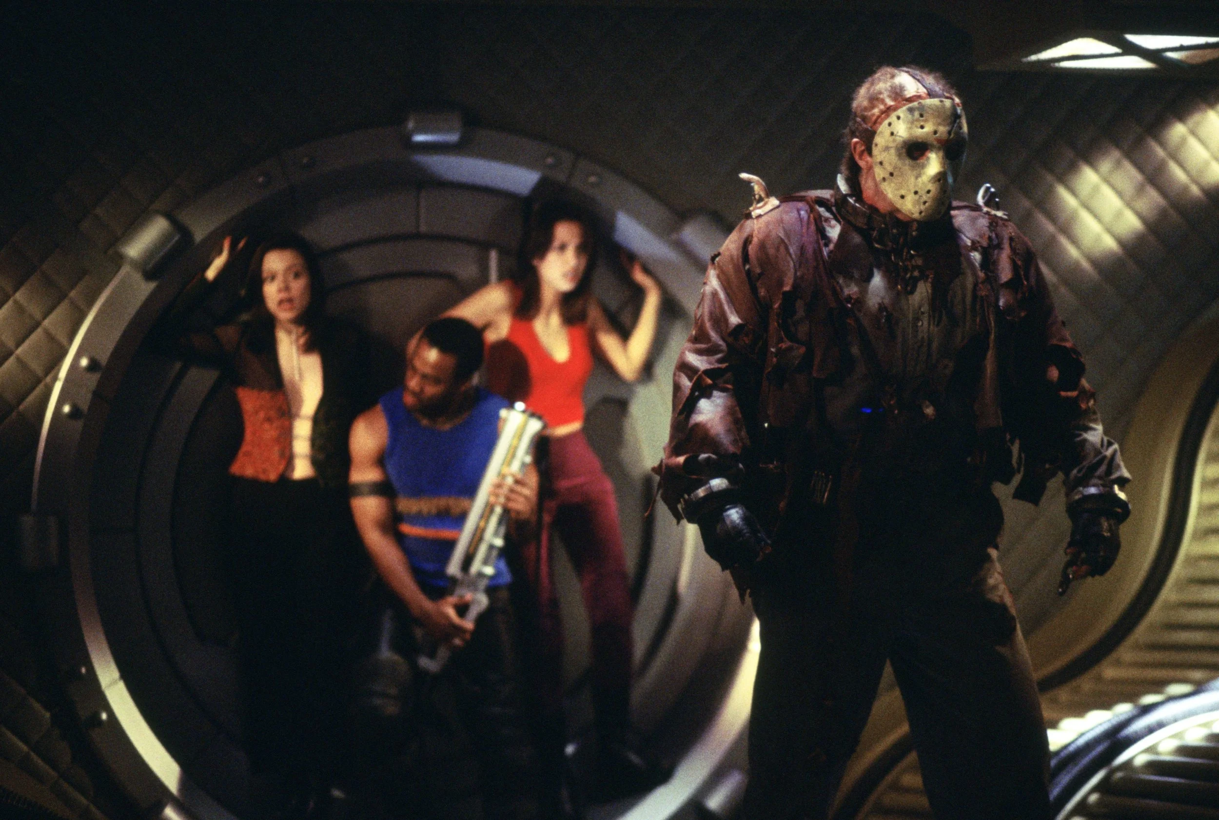 jason-x-2 | film.org.pl Masked figure in a worn hockey mask and brown costume strides toward the camera in a sci‑fi tunnel, as people in colorful outfits watch in the background.