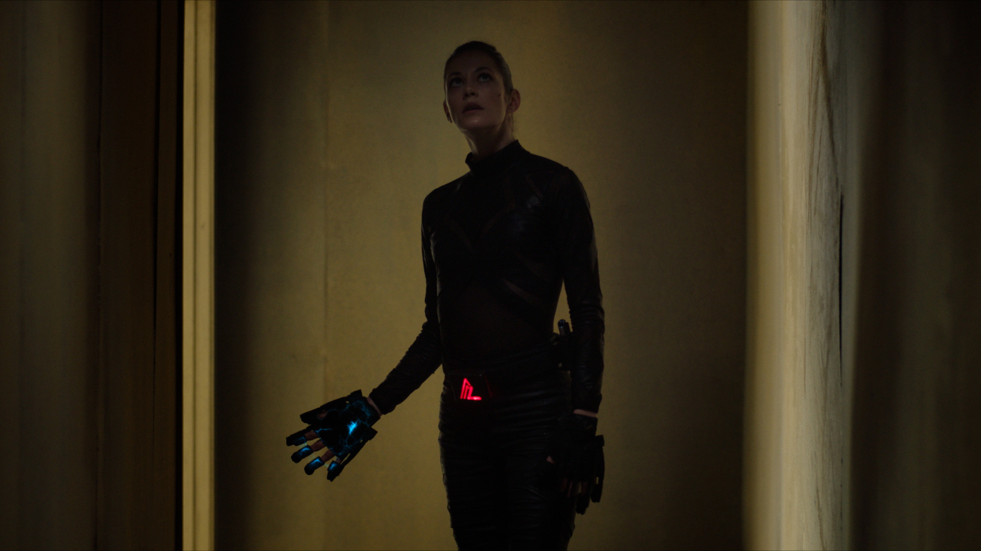 Person in a dark, form-fitting suit stands in a dim hallway; a glowing red emblem sits at the waist and a blue cybernetic glove is on the left hand.