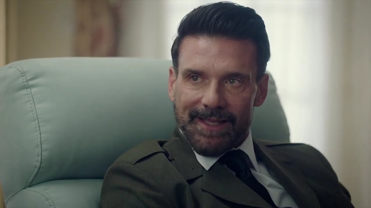 Man in a brown suit and tie sits in a pale green armchair, talking with a slight smile.