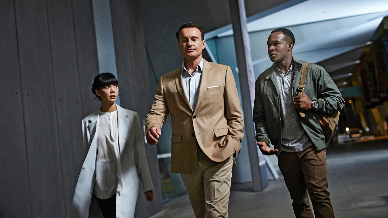Three professionals walk through a modern urban area at night: a man in a tan suit leading, a woman in a light coat beside him, and another man in a green jacket behind.