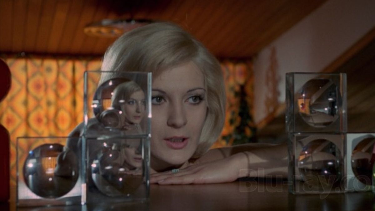 krwawyoboz1971 2 | film.org.pl Blonde woman gazes at glass display blocks with mirrored reflections on a wooden bar surface.