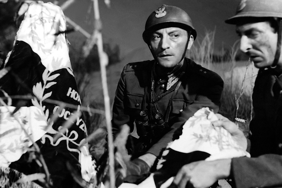 Two soldiers in helmets carry or tend to a wrapped body in a grassy field during a war scene, black-and-white photo.