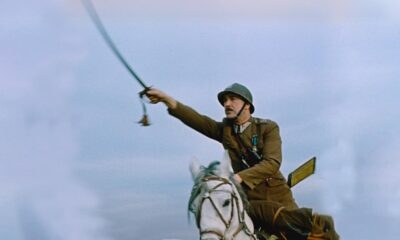 Soldier in a brown uniform riding a white horse, swinging a saber against a pale blue sky.