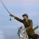 Soldier in a brown uniform riding a white horse, swinging a saber against a pale blue sky.