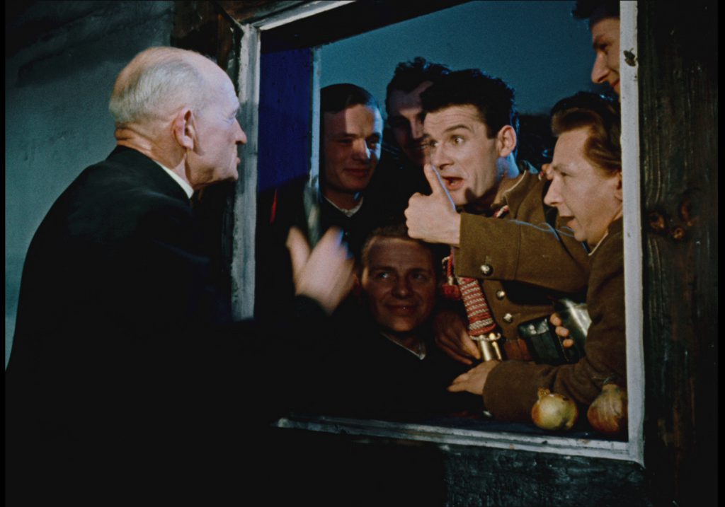 Older man outside a window extends his arm to a crowd of soldiers inside, who cheer and give a thumbs up.
