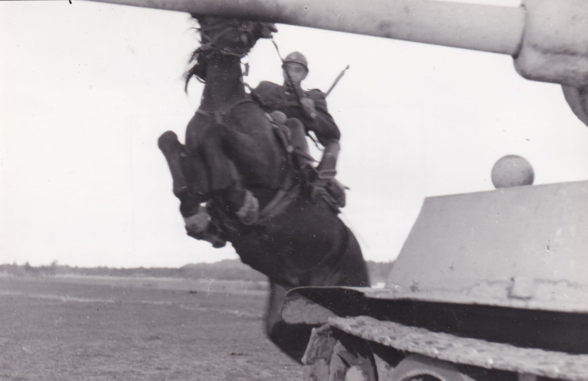 Blurred black-and-white photo of a soldier wearing a helmet, seated on a large metallic structure outdoors; background shows an open space and a rounded object on the right side.