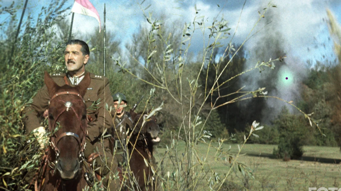 Army officer on a brown horse charging through tall grass, flag waving with smoke in the distance behind him.