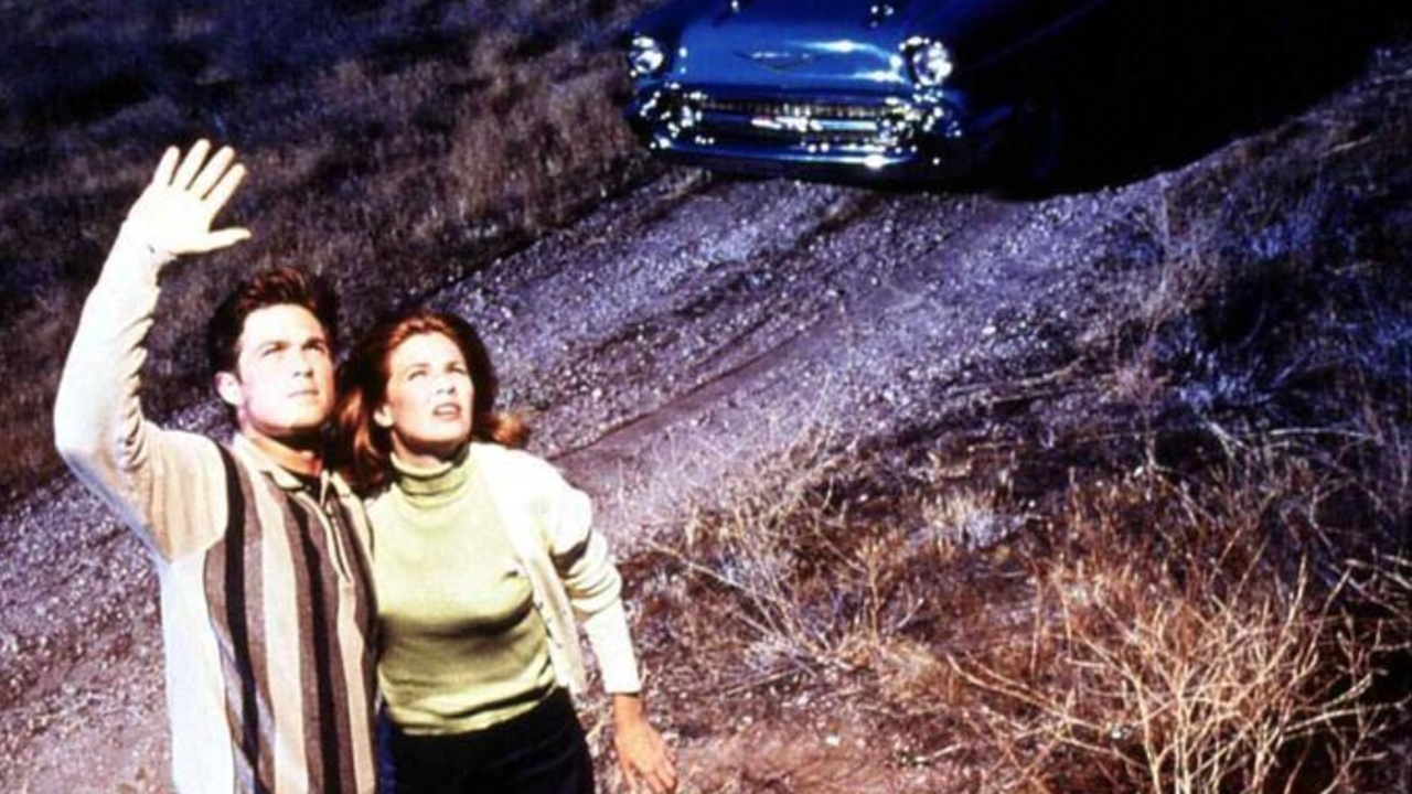 A man and woman stand on a rocky hill, gazing upward at a blue car perched above them, the man raising his hand