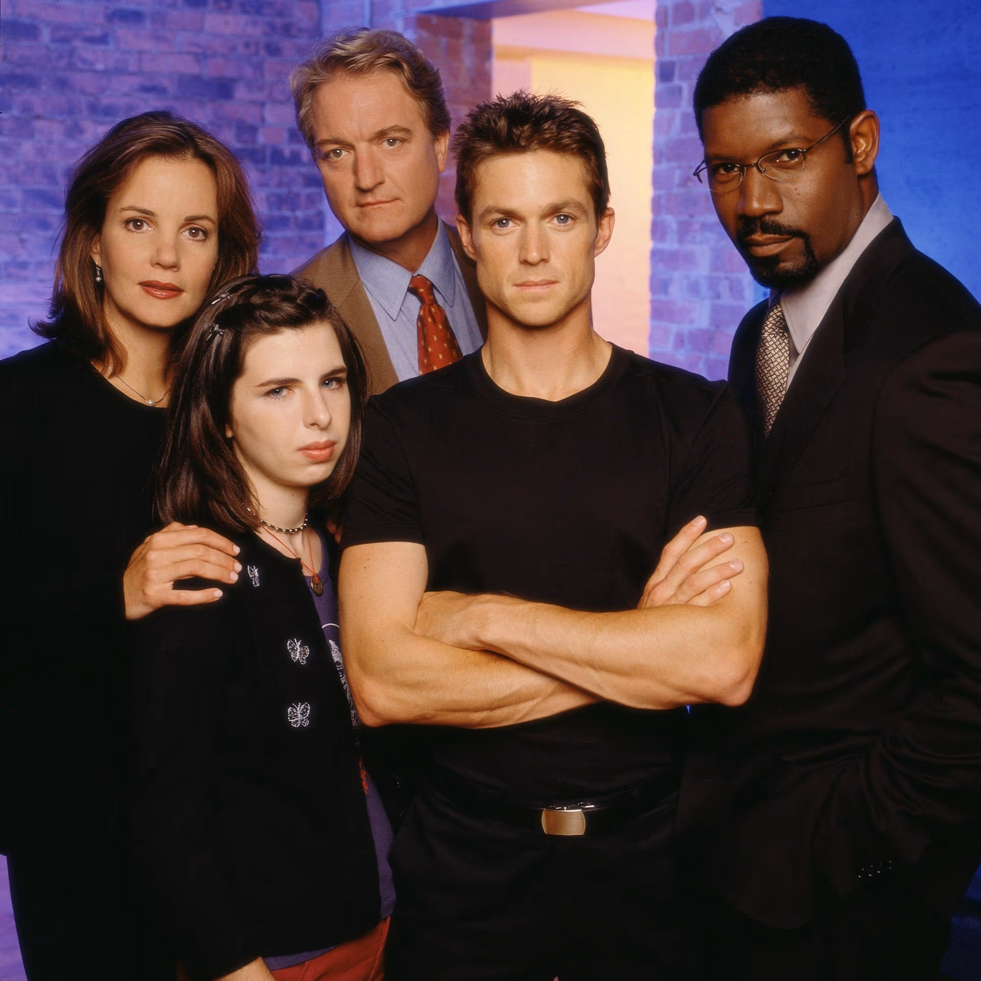 Group portrait of five adults standing close together in a studio with blue-purple lighting and a brick wall background, looking at the camera.