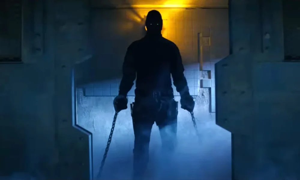 Silhouetted figure in tactical gear with chains in each hand walks through a blue-lit industrial corridor.