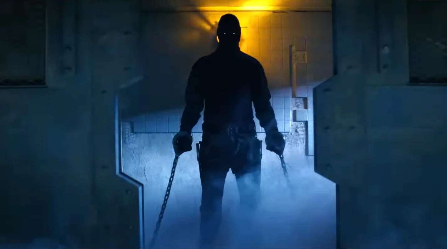 Silhouetted figure in tactical gear with chains in each hand walks through a blue-lit industrial corridor.