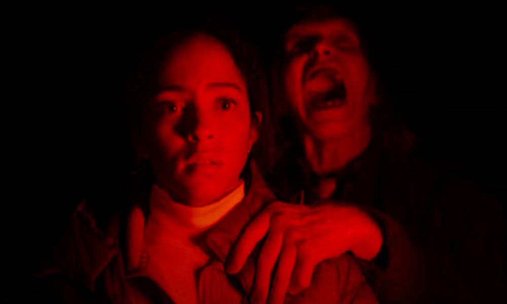 Two people in red light huddle together at night; the front person looks startled while the person behind screams.
