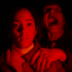 Two people in red light huddle together at night; the front person looks startled while the person behind screams.