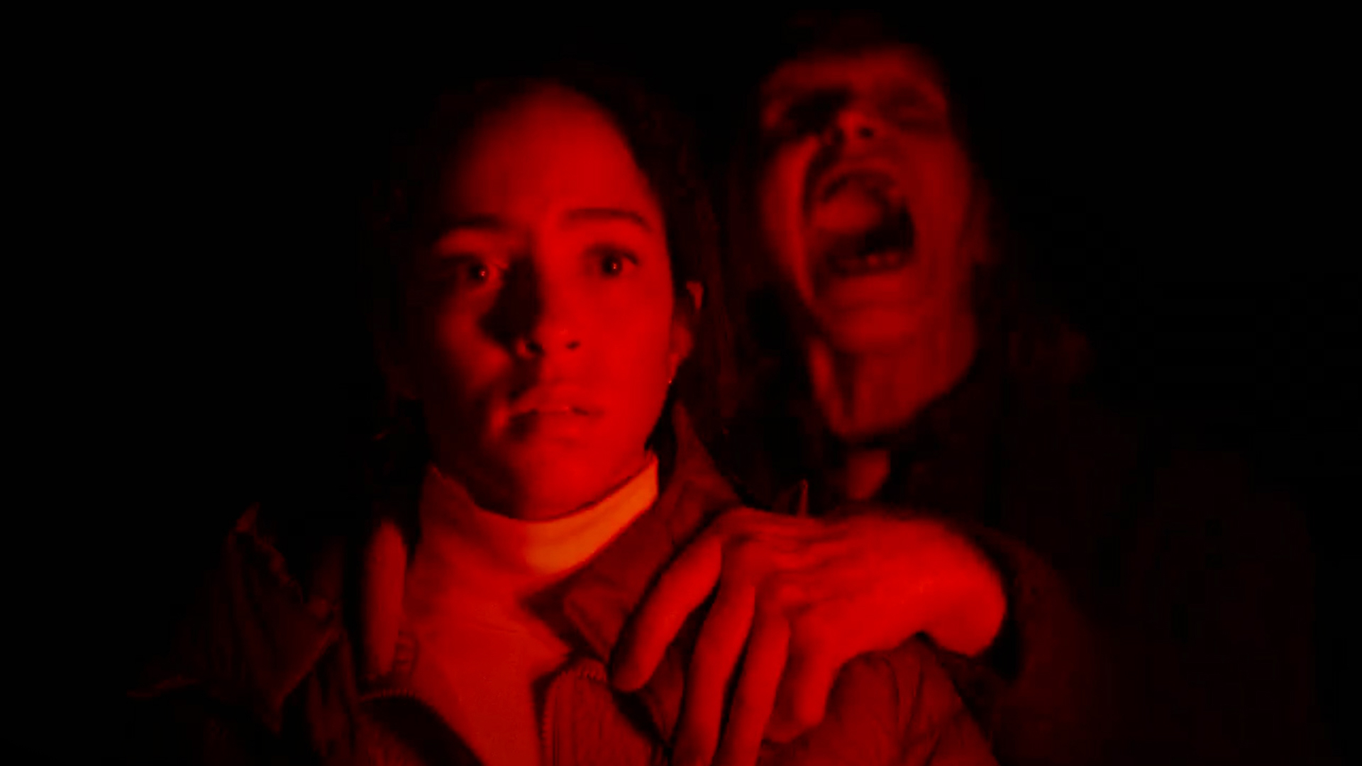 Two people in red light huddle together at night; the front person looks startled while the person behind screams.