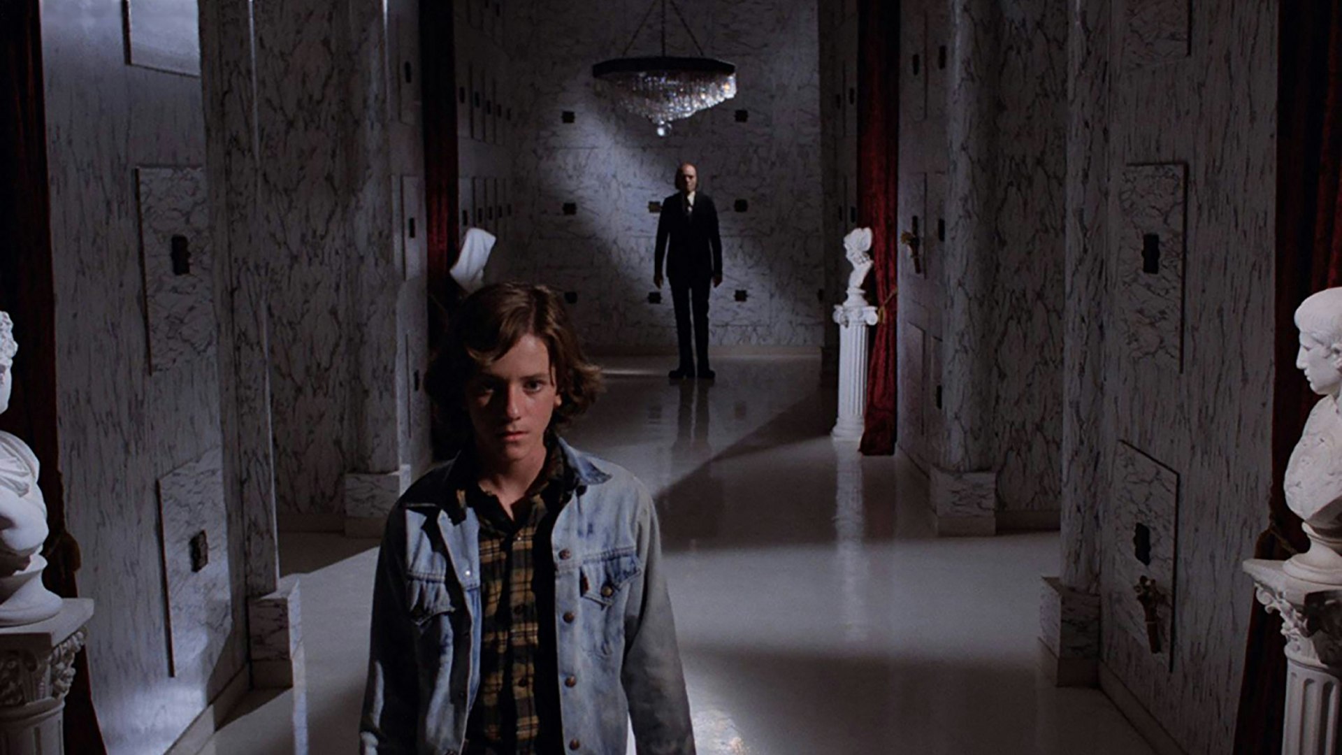 Young man in a denim jacket stands in a marble hallway lined with busts; a man in a suit appears in the distance beneath a chandelier.