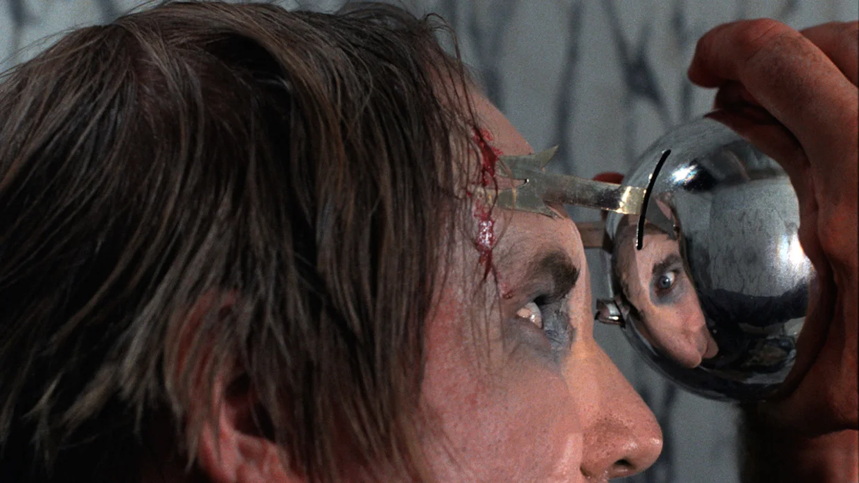 Man with a bleeding forehead looks into a handheld circular mirror held by another person.