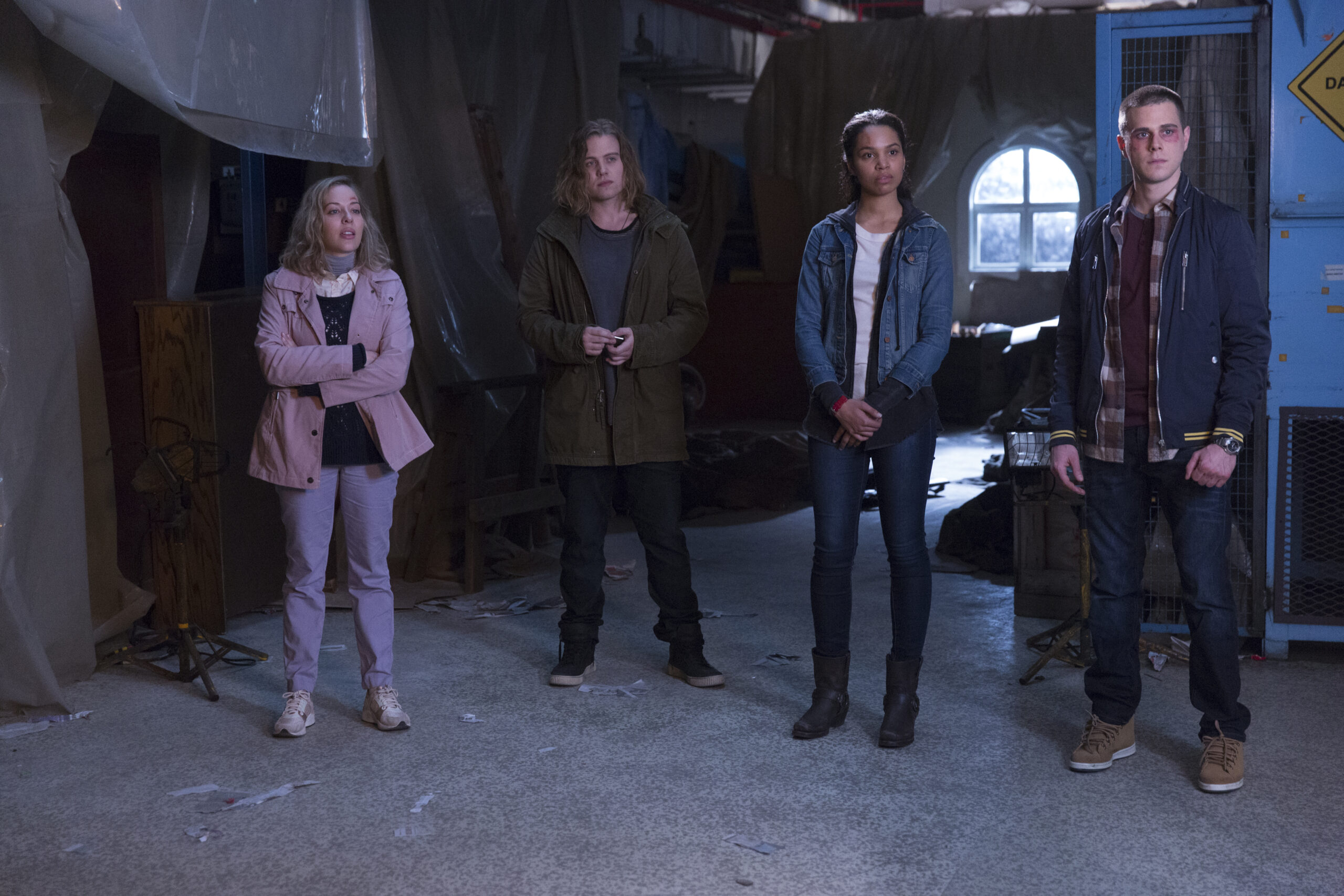 Four young adults stand in a dim, cluttered warehouse-like space, facing the camera: woman in pink jacket, man in olive parka, woman in denim jacket, man in a blue jacket and plaid shirt.