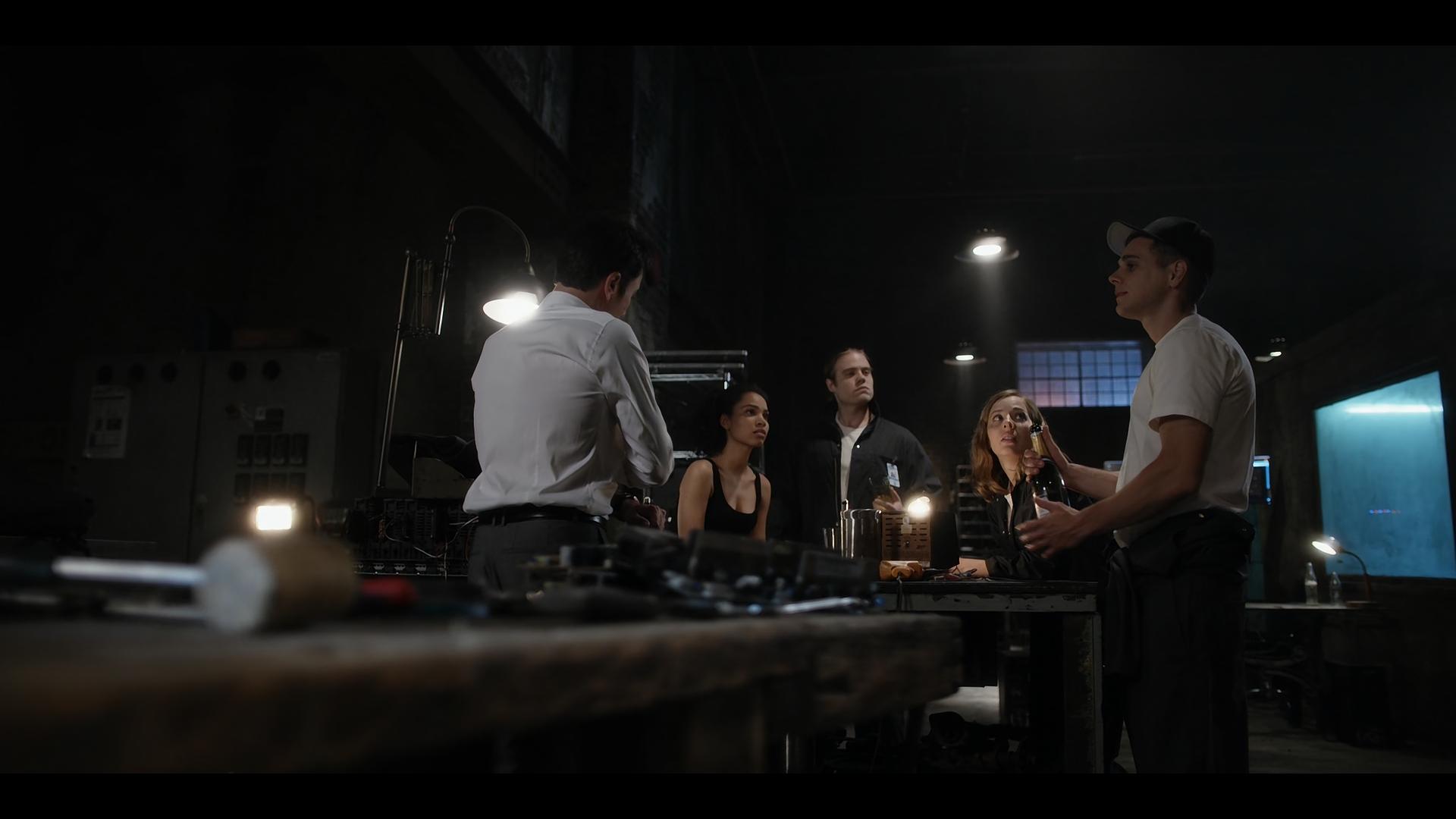 Five people stand and sit around a cluttered workbench in a dark industrial room, one person holding a bottle under overhead light.