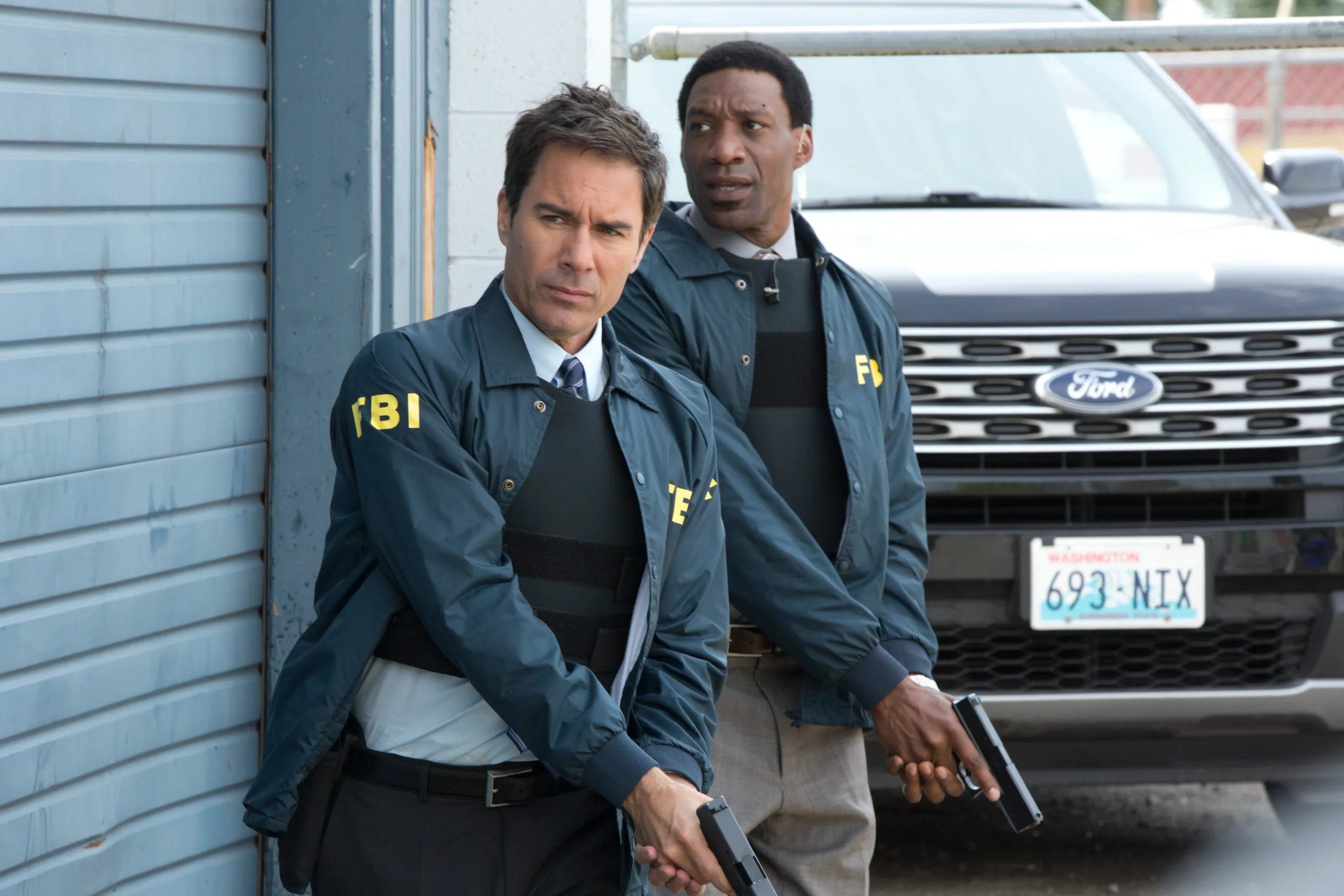 Two FBI agents in dark jackets with yellow FBI lettering stand near a blue building, each holding a handgun and looking alert.