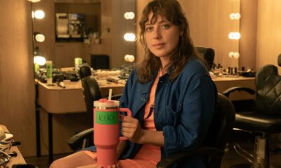 Young woman in a makeup room sits in a wheelchair, holding a pink tumbler labeled 'KIKI' with vanity lights in the background.