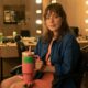 Young woman in a makeup room sits in a wheelchair, holding a pink tumbler labeled 'KIKI' with vanity lights in the background.