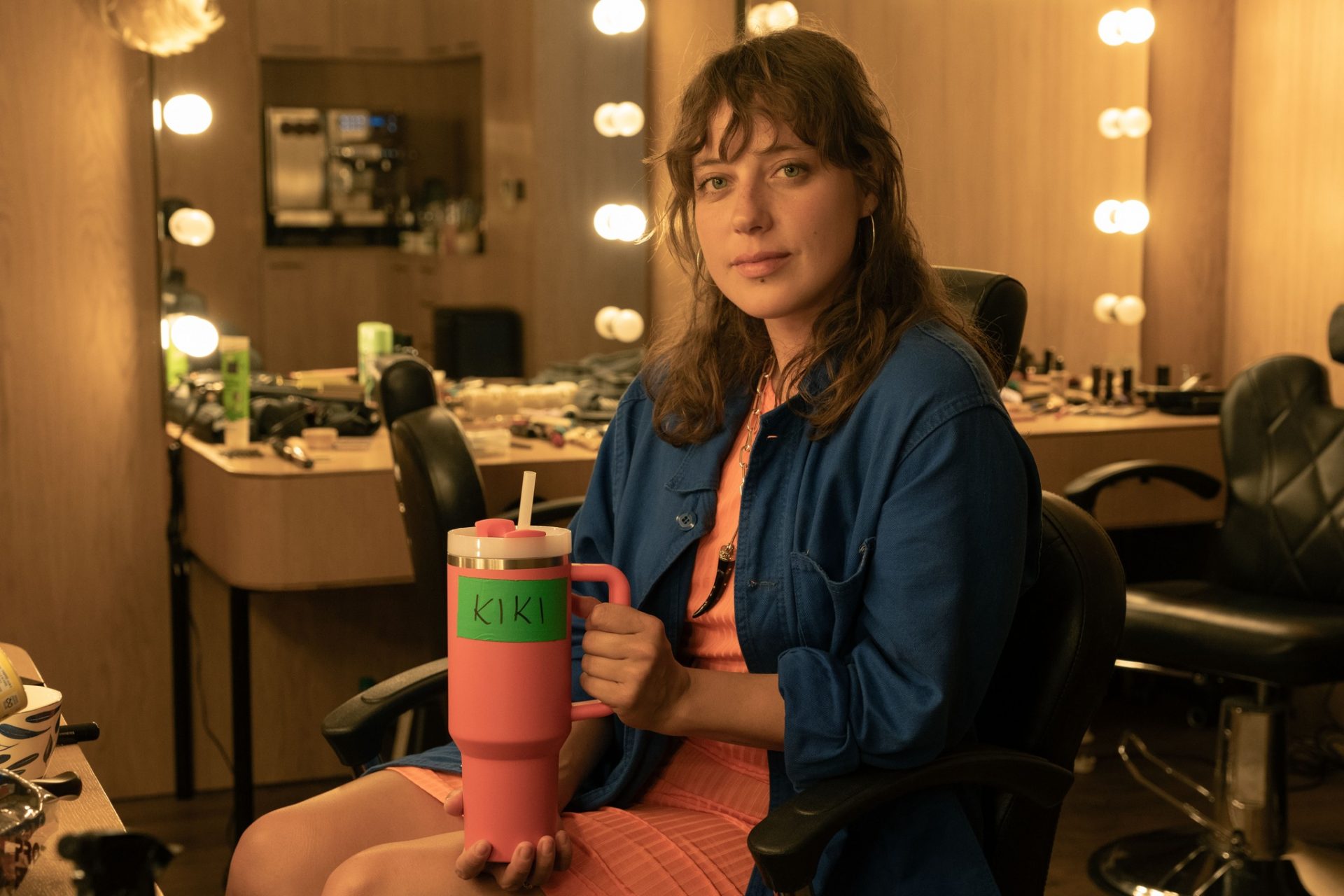proud-4 | film.org.pl Young woman in a makeup room sits in a wheelchair, holding a pink tumbler labeled 'KIKI' with vanity lights in the background.