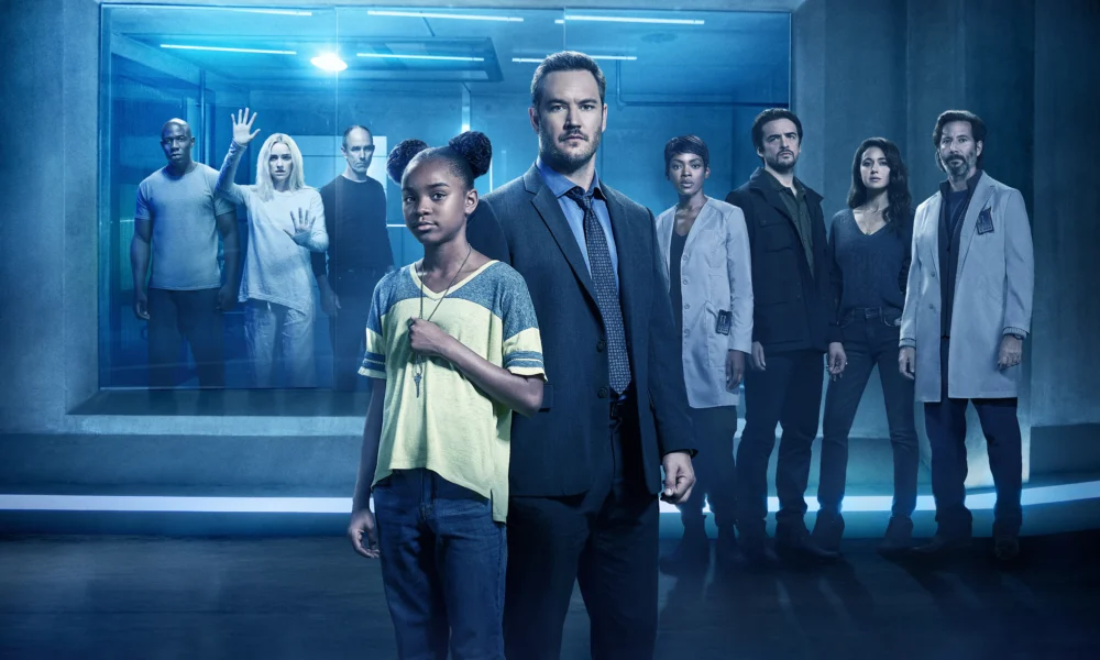 Diverse group of professionals standing in a blue-lit, futuristic corridor, some in lab coats and one in a suit, facing the camera.