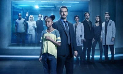 Diverse group of professionals standing in a blue-lit, futuristic corridor, some in lab coats and one in a suit, facing the camera.
